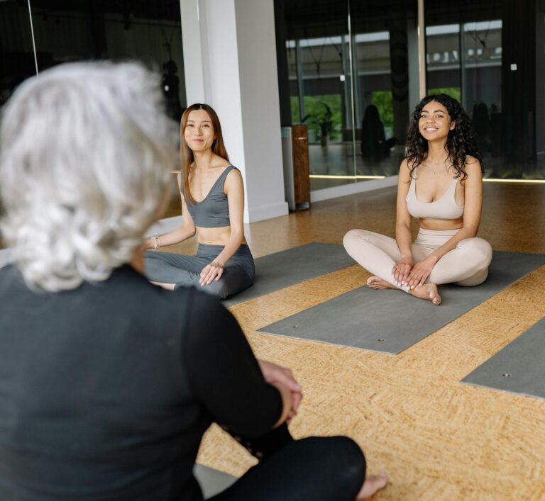 Two women and an instructor in a yoga class practicing poses on mats inside a modern studio.
